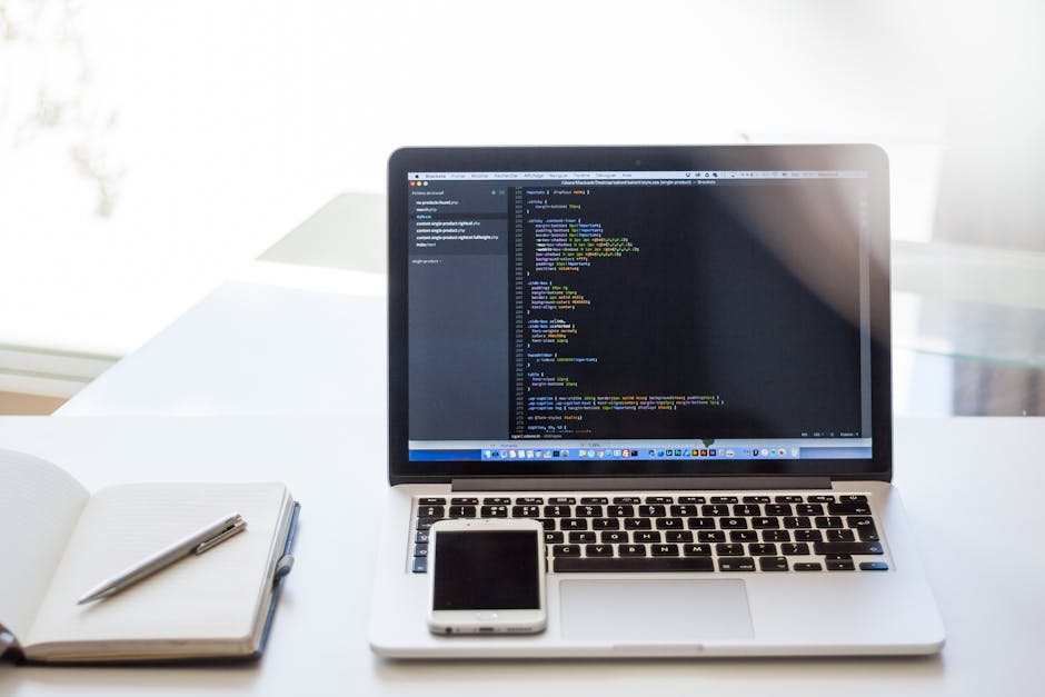 A close-up view of a modern workspace featuring a silver laptop with an open screen displaying lines of computer code on a dark background, indicating programming or software development activity. The